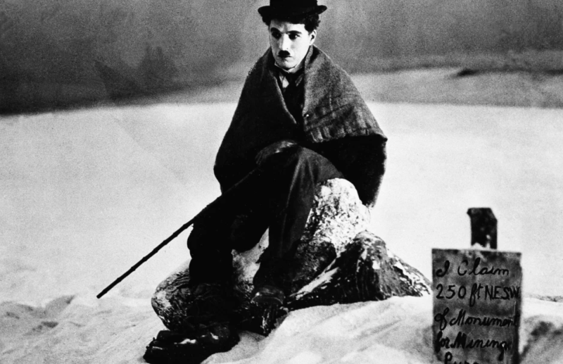 A white man with a square mustache sits on a snowy bank wearing a bowler hat and blanket around his shoulders. A walking stick is at his side.