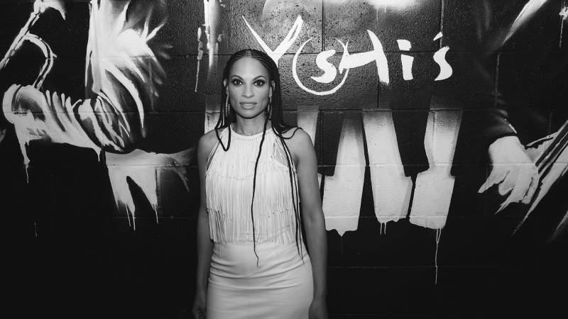 An African American woman in braids and a white dress poses in front of a wall with paint stencils on it.