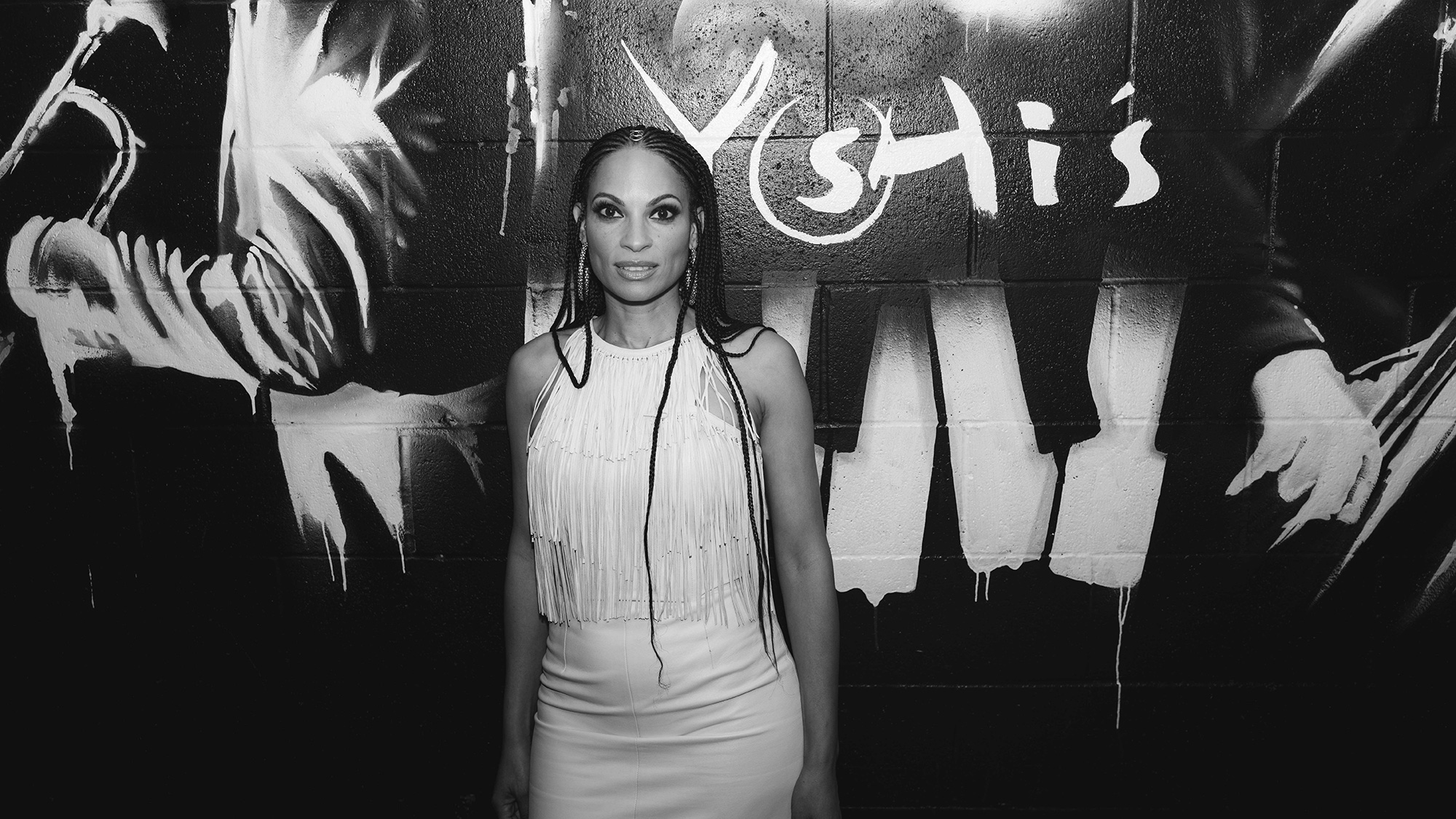 An African American woman in braids and a white dress poses in front of a wall with paint stencils on it.