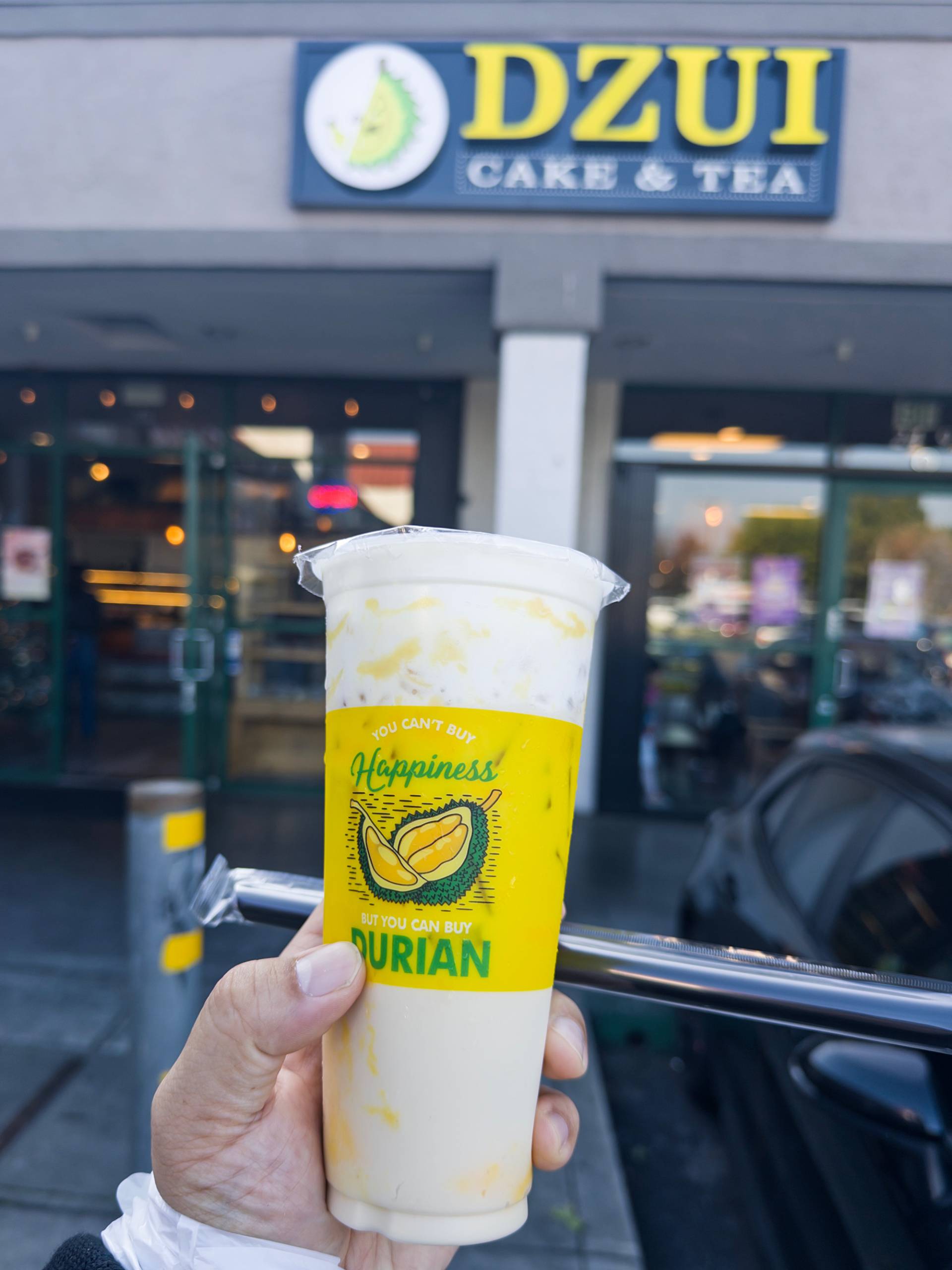 Hand holding a cup of milk tea in front of Dzui Cake & Tea. The text on the cup reads, "You can't buy happiness, but you can buy durian."