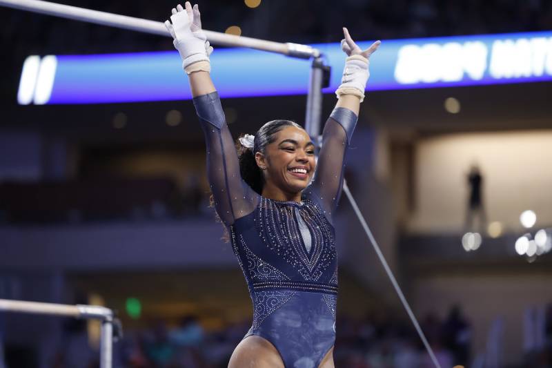 A young Black gymnast wearing a dark blue leotard, smiles and raises her arms straight over her head at the end of a routine. Her hair is tied back in a neat ponytail.