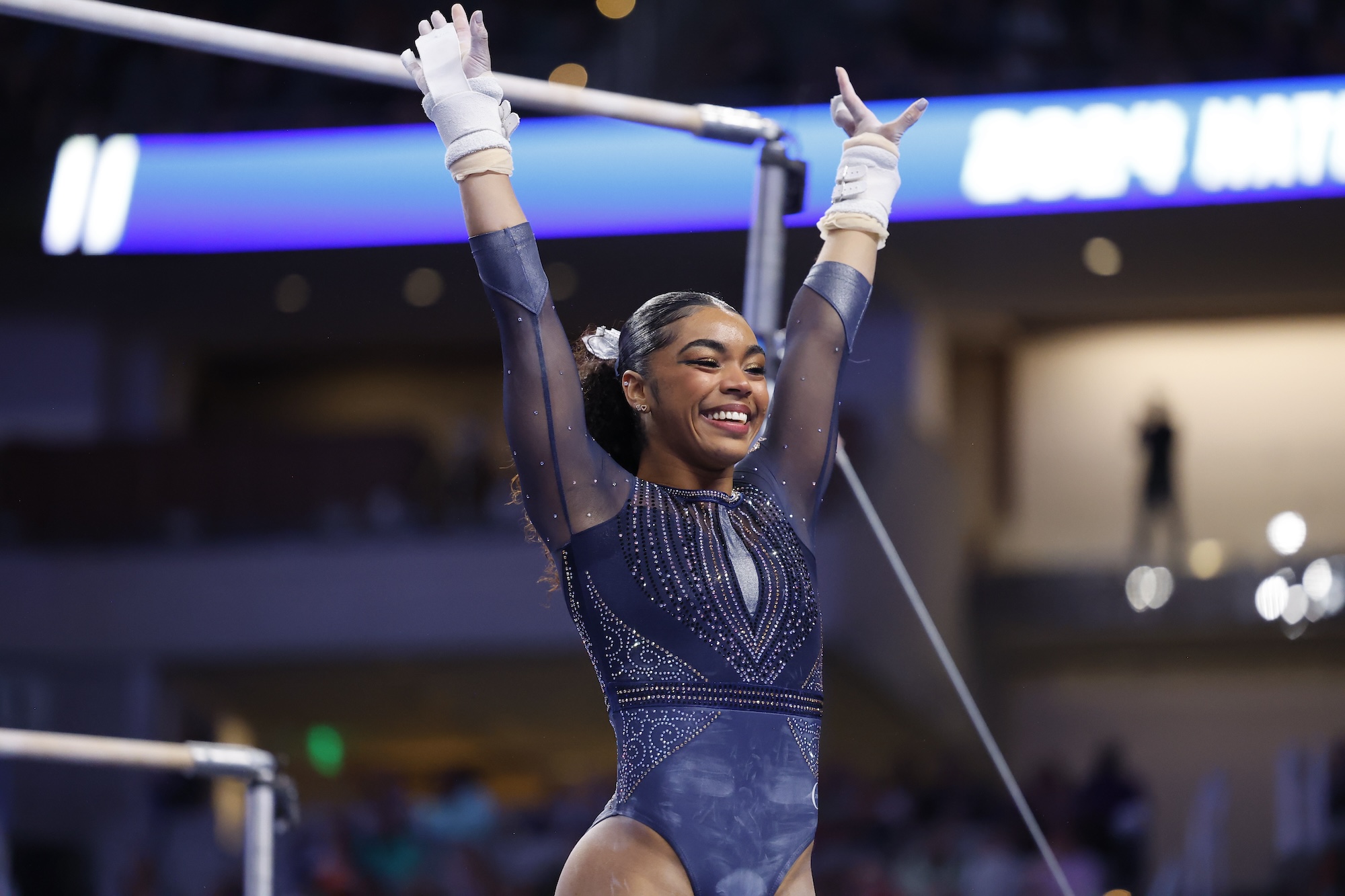 A young Black gymnast wearing a dark blue leotard, smiles and raises her arms straight over her head at the end of a routine. Her hair is tied back in a neat ponytail.