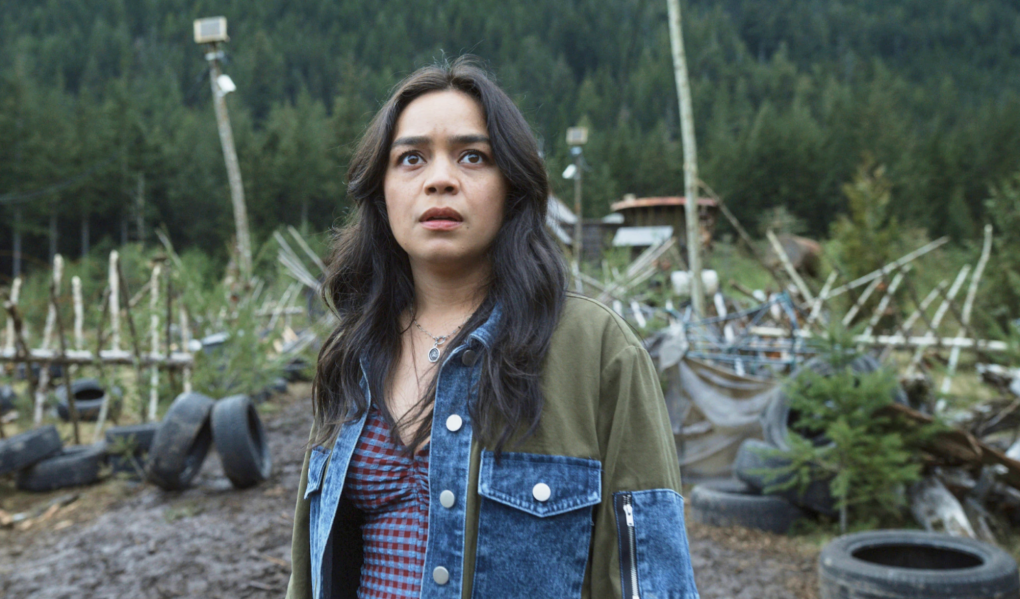 A young, mixed race woman with long brown hair, wearing a denim shirt, stands in a lot full of spiky junk, looking into the distance, horrified.