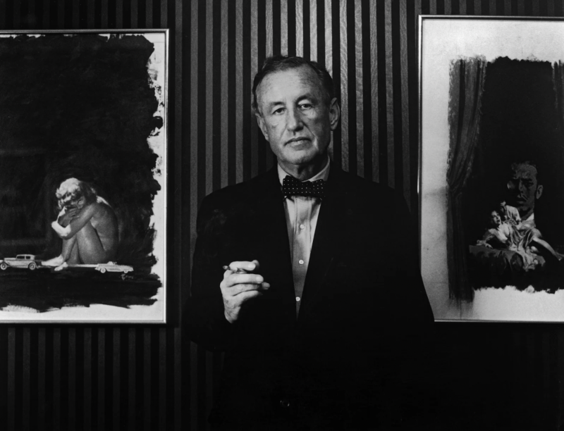 An older white man wearing a smart suit and bowtie stands, casually smoking against a striped wall, paintings hanging either side of him.