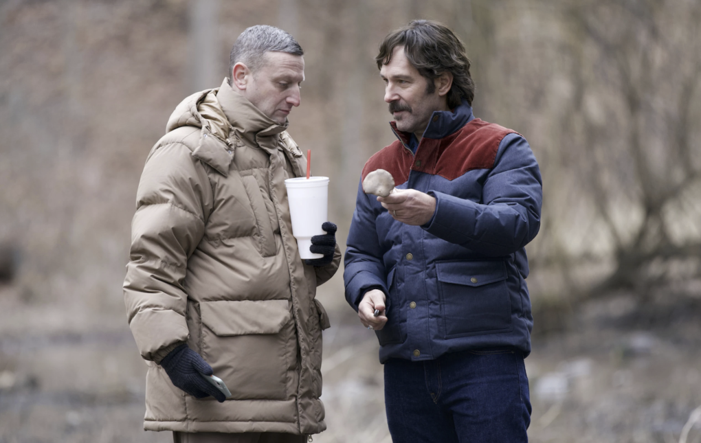 Two middle aged men wearing puffy winter coats stand together in the woods.