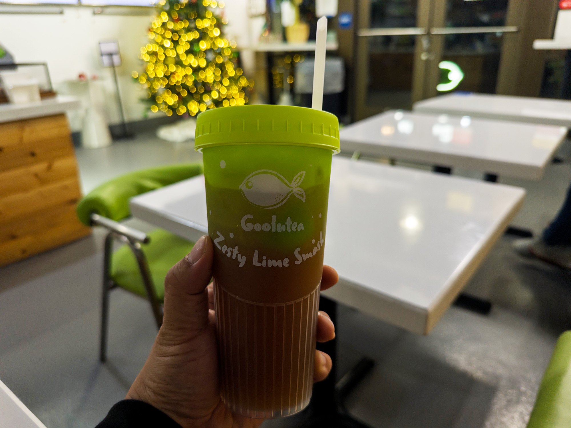 Hand holding a boba drink with a Christmas tree in the background. The text on the cup reads, "Goolutea Zesty Lime Smash"