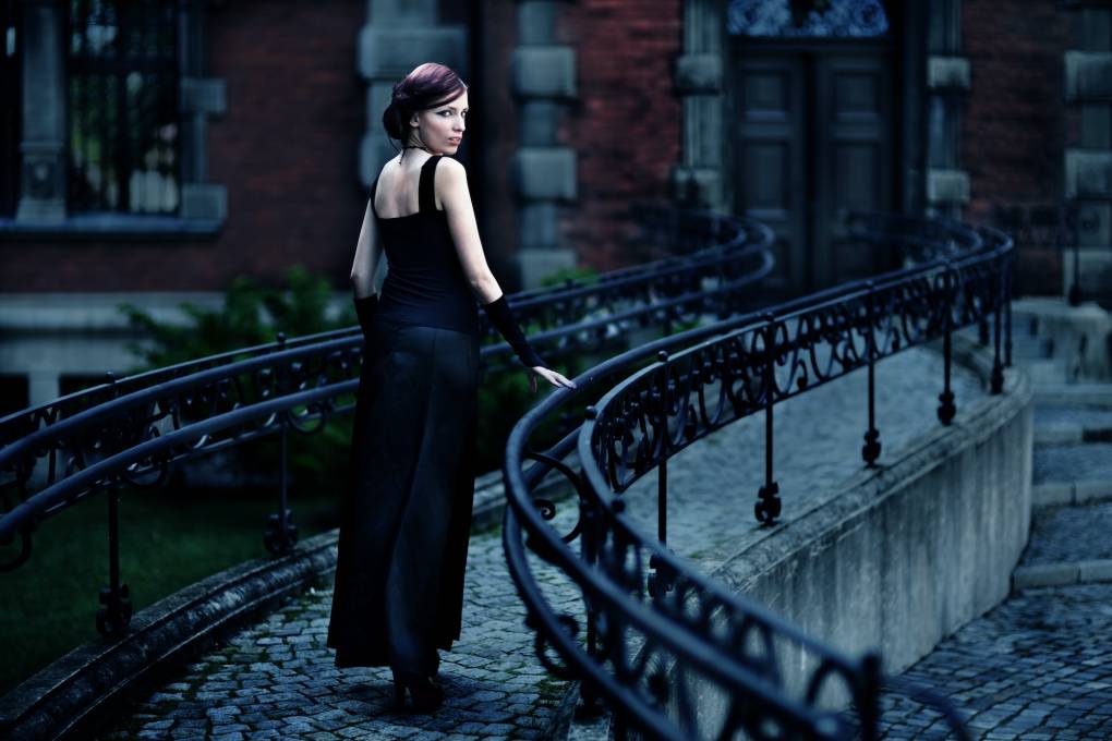 A pale white woman with purple hair in a long black dress walks along a narrow path with elaborate iron railings.