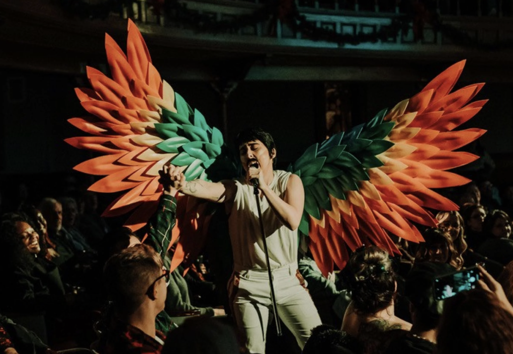 A drag king on stage, singing while wearing dirty white jeans and a t-shirt with huge orange and green wings.