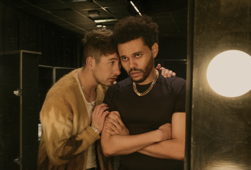 A white man wearing a beige cardigan and white t-shirt embraces a Black man wearing a black t-shirt who is standing arms folded, with tears in his eyes, looking into a dressing room mirror.
