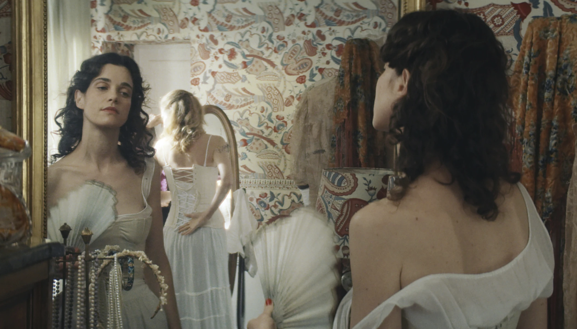 A woman in an off the shoulder white gown with bodice examines her reflection in a mirror. Another woman behind her is also trying on a white gown with corset.