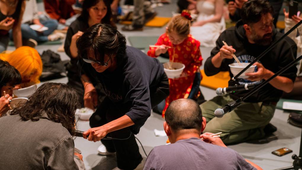 Several people crouch on the ground slurping udon noodles while a man captures the sound with a microphone.