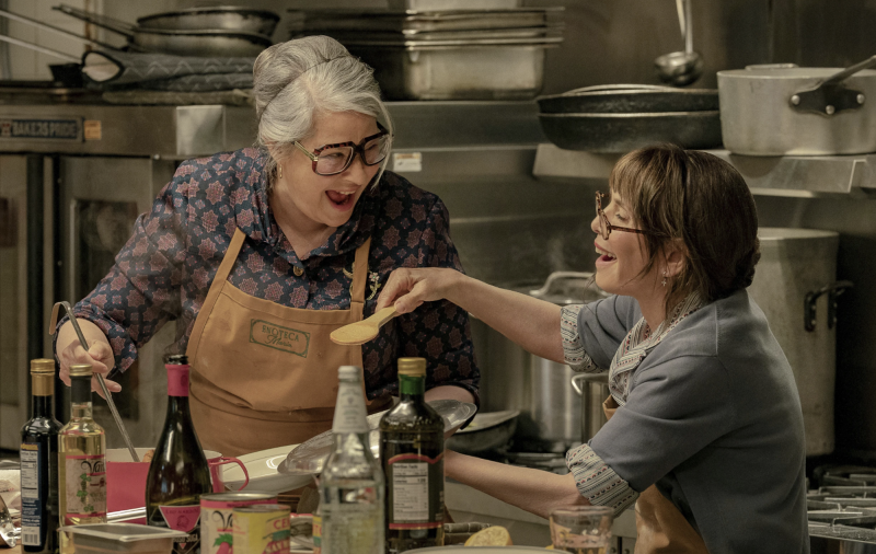 Two laughing women with grey hair, wearing brown aprons, taste a dish inside a restaurant kitchen.