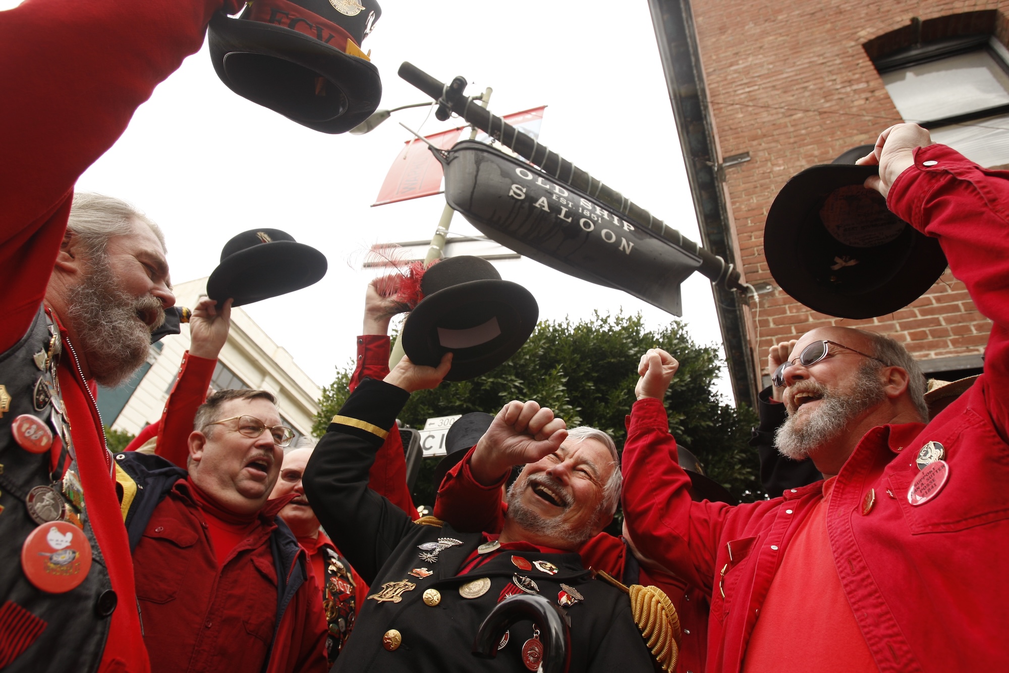 A group of older men stand cheering underneath an outdoor bar sign at the Old Ship Saloon. They are raising their arms and cheering.