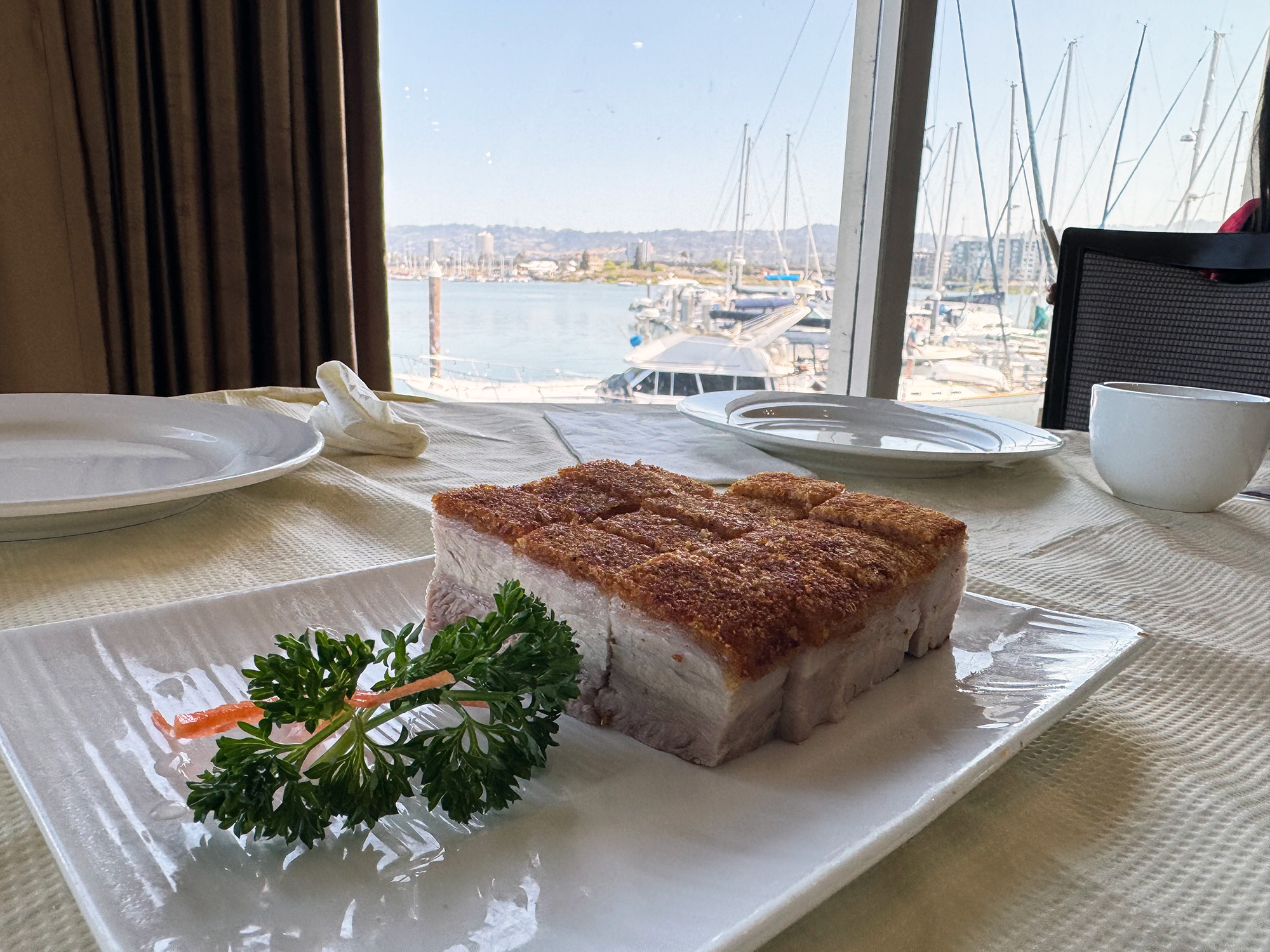 Crispy-skinned roast pork belly on a table in front of a window showing views of yachts in the marina.
