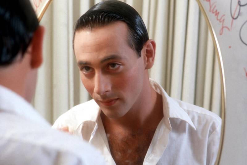 A white man with slicked back black hair looks at his face in a mirror, wearing an open white shirt.