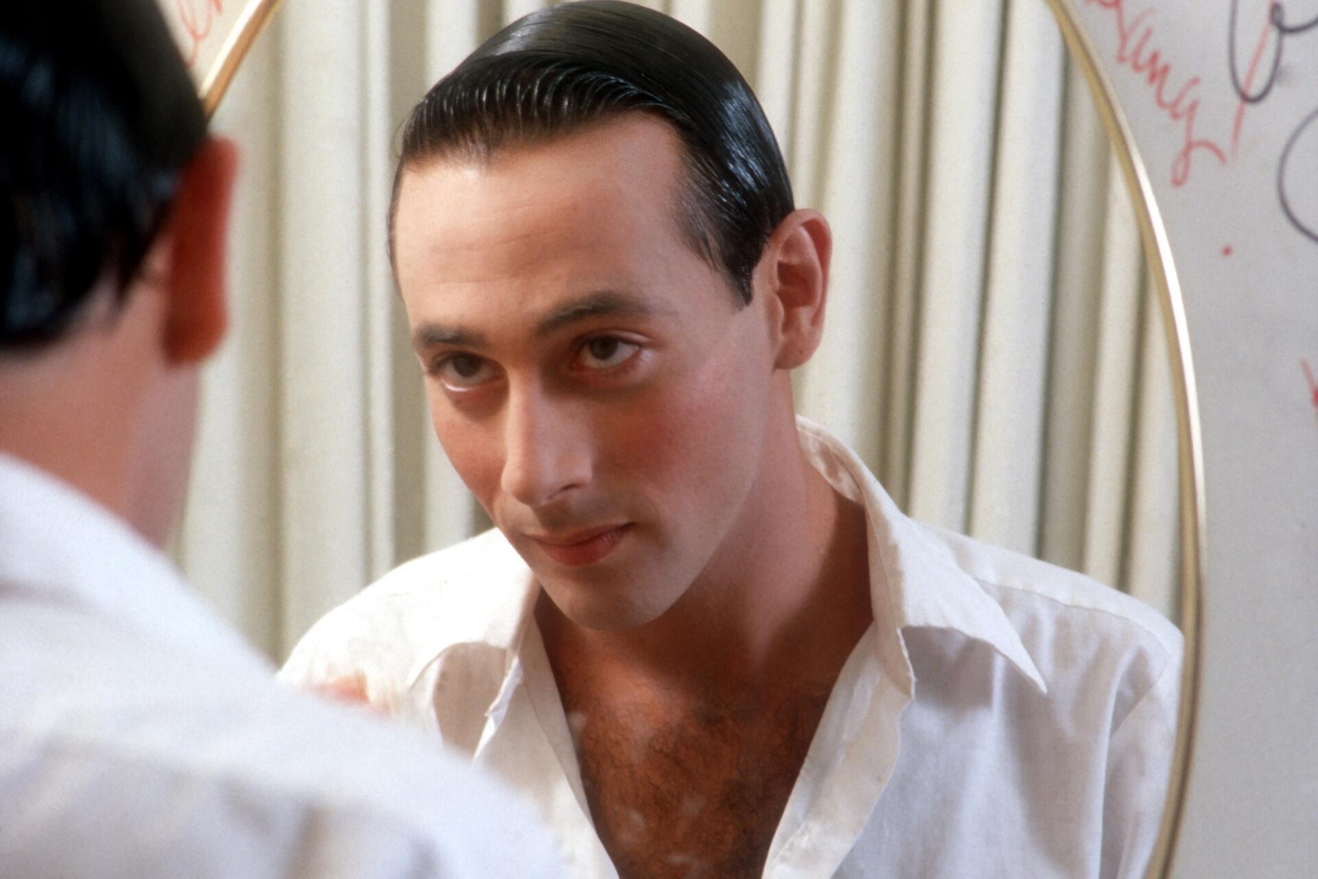 A white man with slicked back black hair looks at his face in a mirror, wearing an open white shirt.