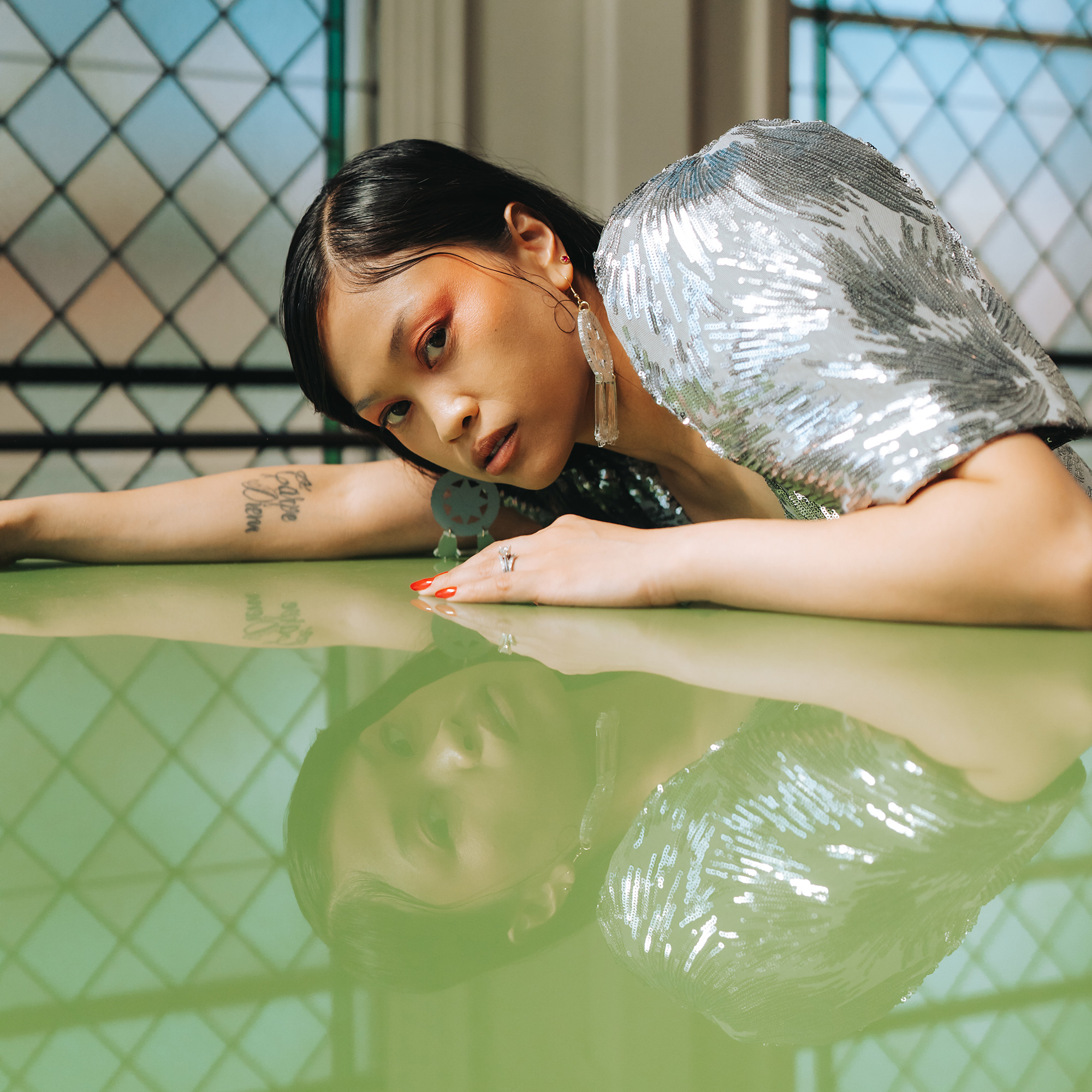 A young Filipina woman in a silver top leans over a shiny green surface, her reflection visible