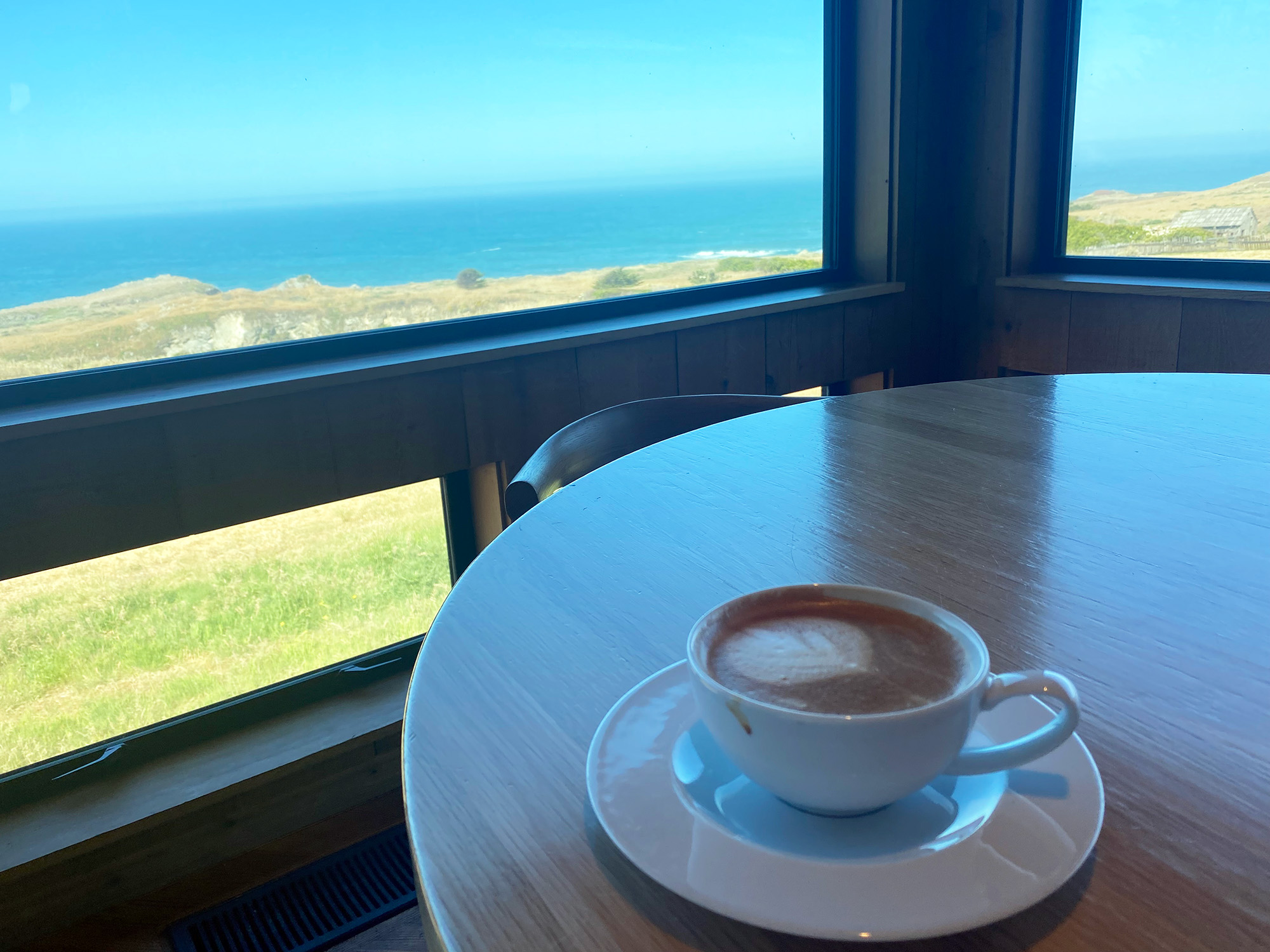 Cappuccino in a white cup on a wood table. The view outside the window is of a grassy cliff and, beyond that, a blue sky and sea.