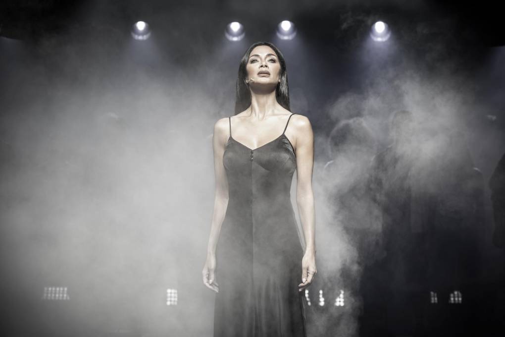 A beautiful mixed race woman wearing a long black negligee stands on stage under spotlights, surrounded by dry ice.