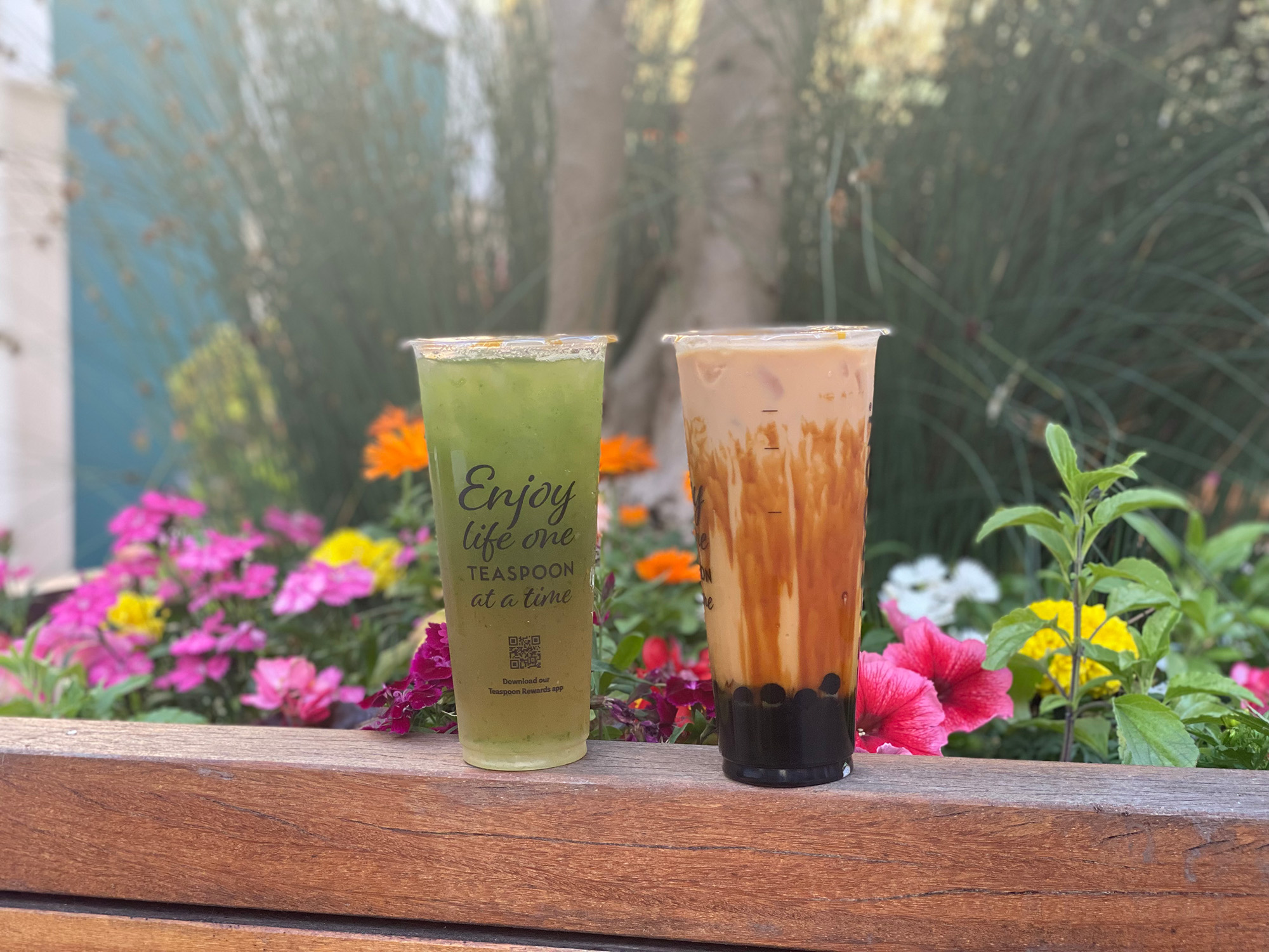 Two boba drinks placed on the edge of a planter box filled with colorful flowers.