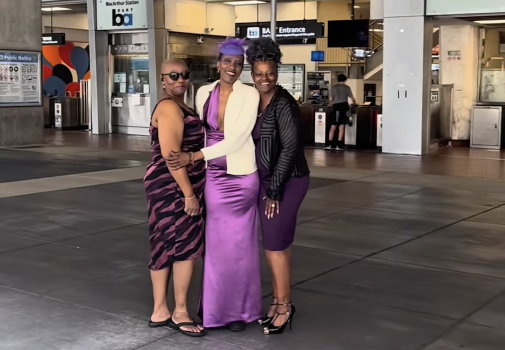 A Black woman in a purple satin gown, purple hat and white suit jacket stands outside a train station, smiling with two friends.
