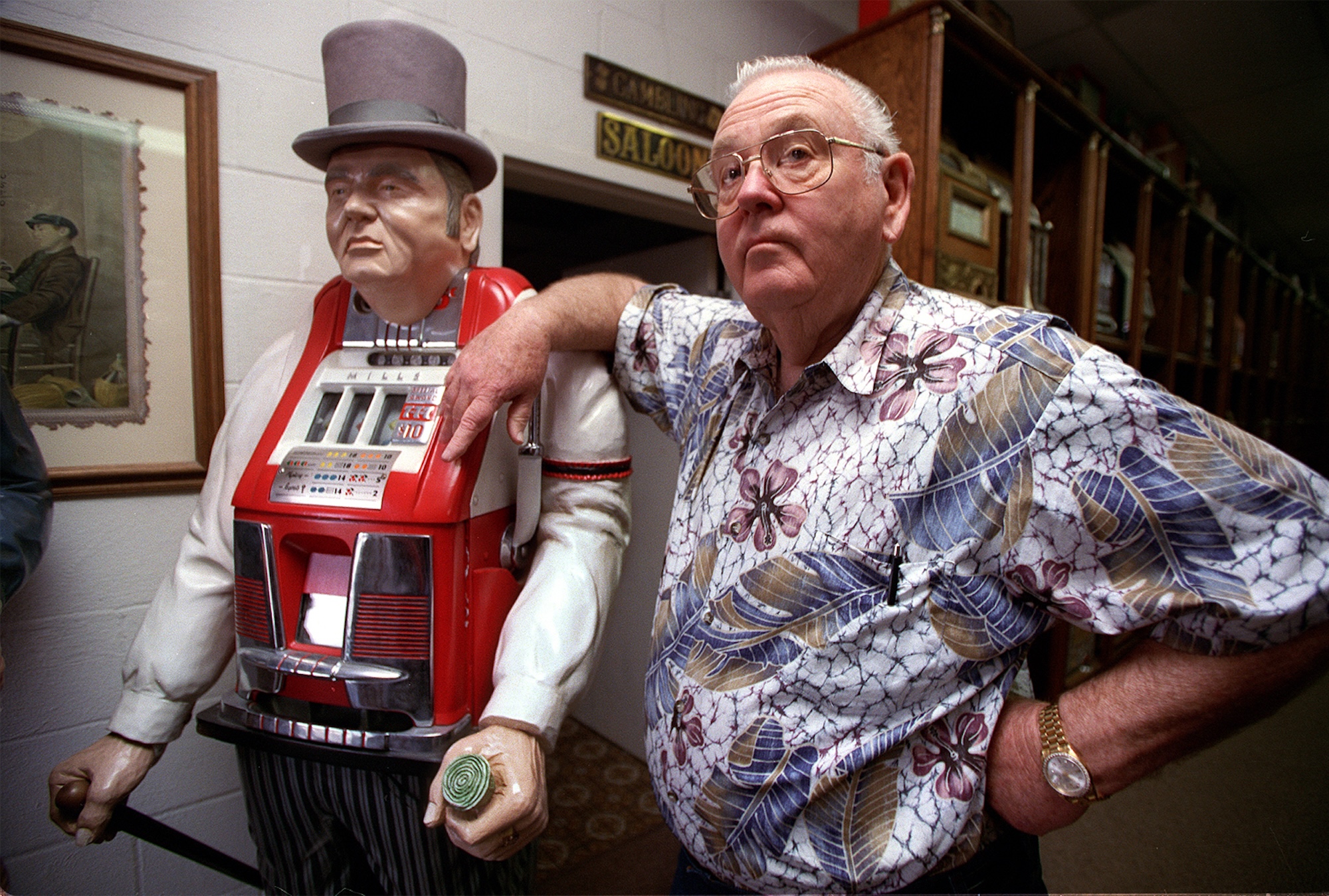 An older gentleman wearing a casual and colorful shirt leans on the statue of a man in a top hat that is also an antique slot machine.