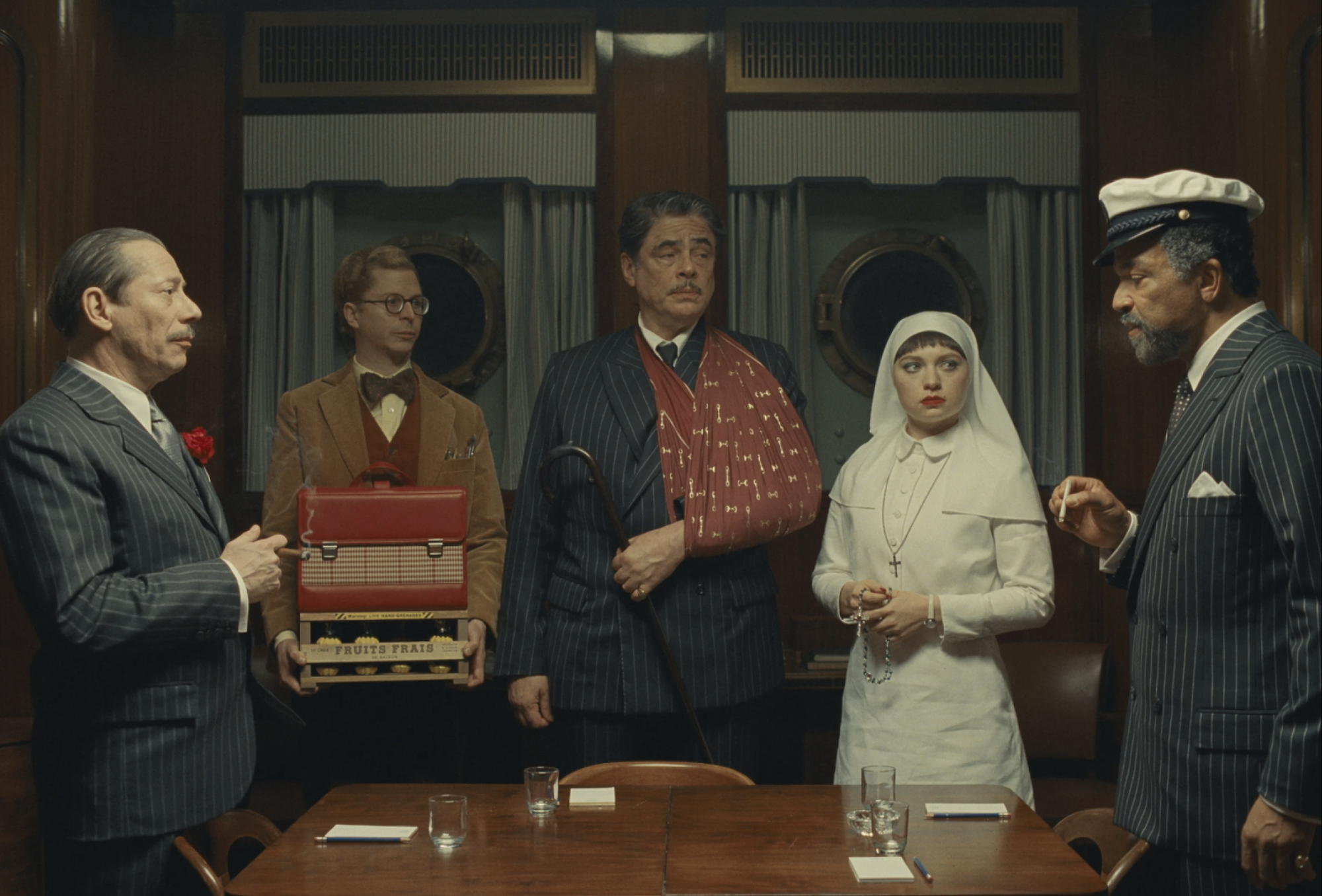 Four men in a variety of old fashioned suits stand with a young nun dressed in a white habit, around an antique table, with blank expressions on their faces.
