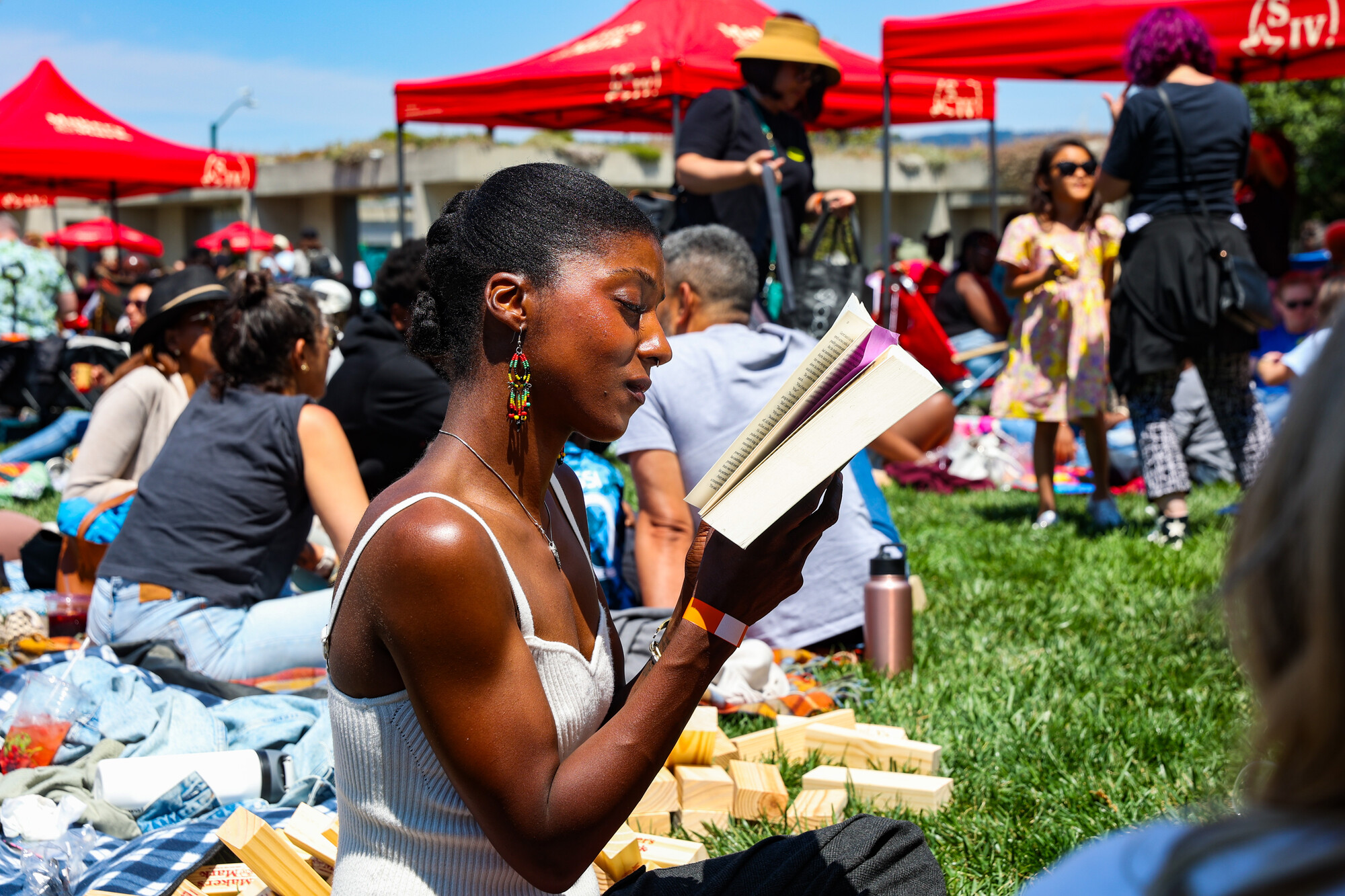 A Black woman reading a book while seated on the grass surrounded by other picnickers.