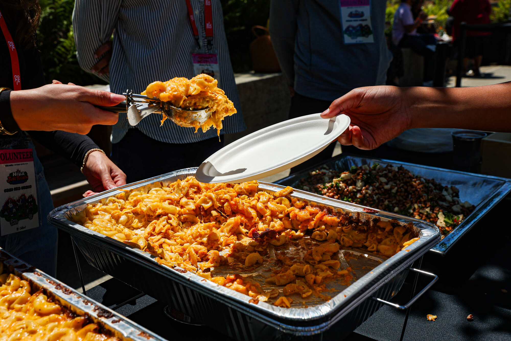 A volunteer ladles a big scoop of mac and cheese from a tray onto a paper plate.