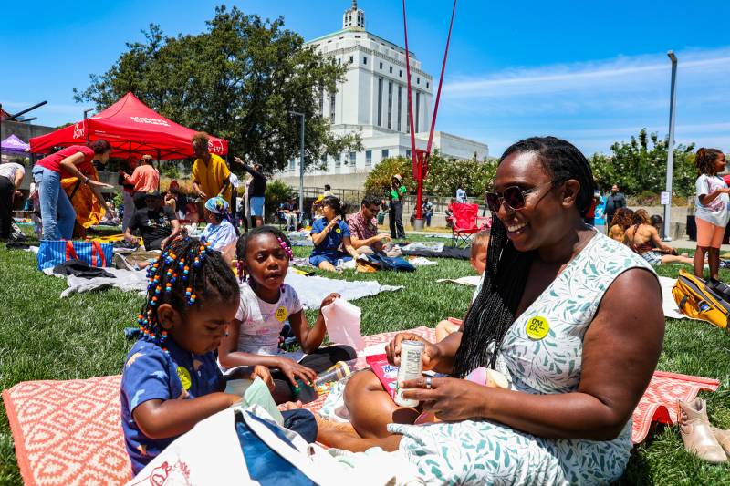 woman with two children smiles and snacks on lawn with Oakland Court House behind