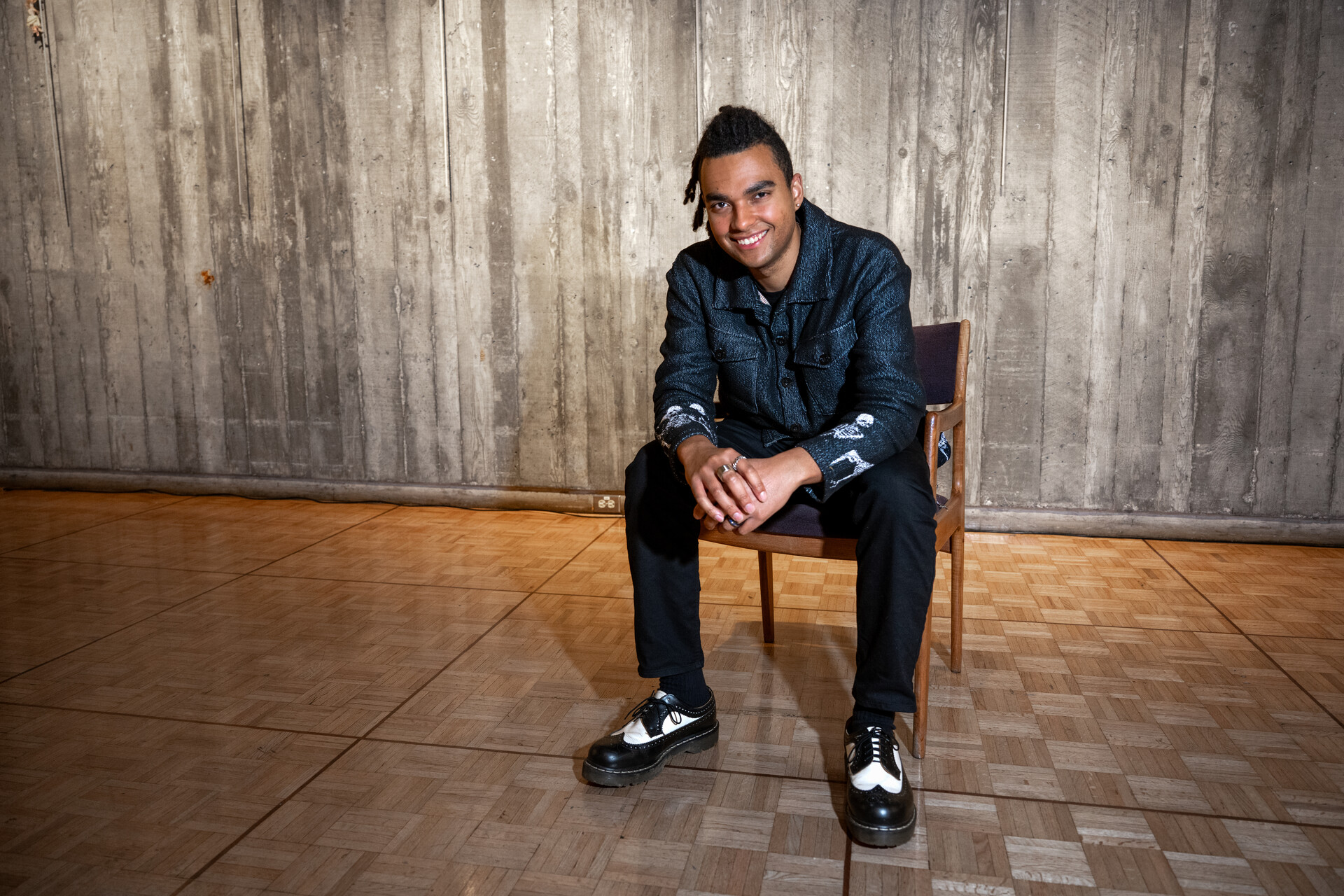A young Black man in a blue shirt and black slacks sits on a wooden chair against a concrete wall, looking into the camera and smiling
