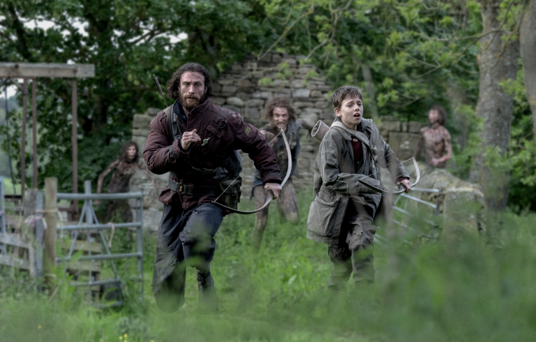 A white man carrying a bow runs through a field, arrows strapped to his back, looking fearful. To his right is a young teenage boy, also desperately running. Behind them, three bloodied and dirtied figures give chase.