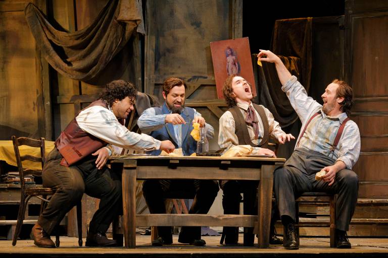 Four men in 1800s clothes frolic and laugh at an old wooden table