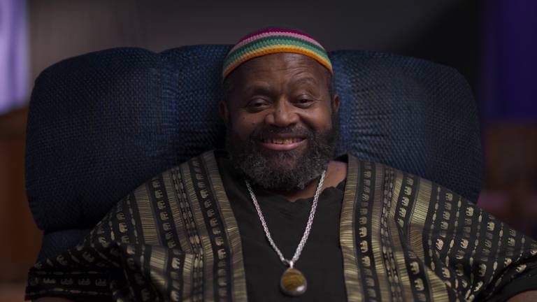 Black man in rainbow beanie sits with big smile
