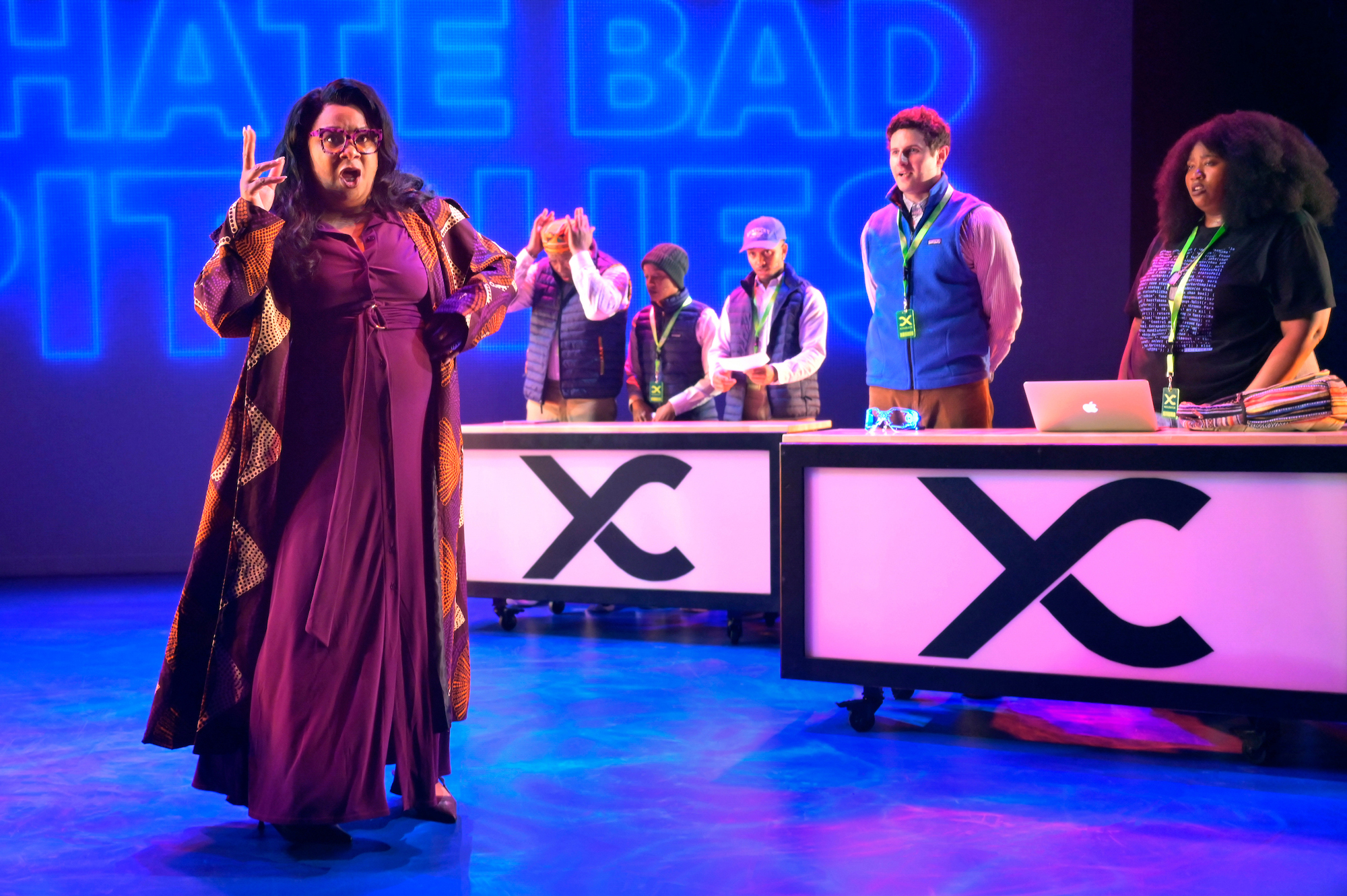 Black woman in foreground with other cast members behind desks
