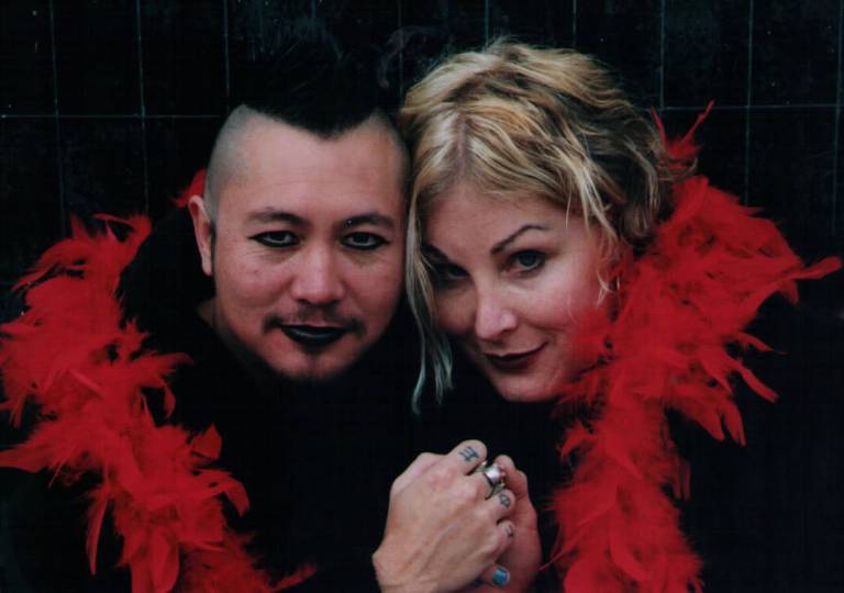 person with dark makeup and person with blonde hair clasp hands, encircled by red feather boa