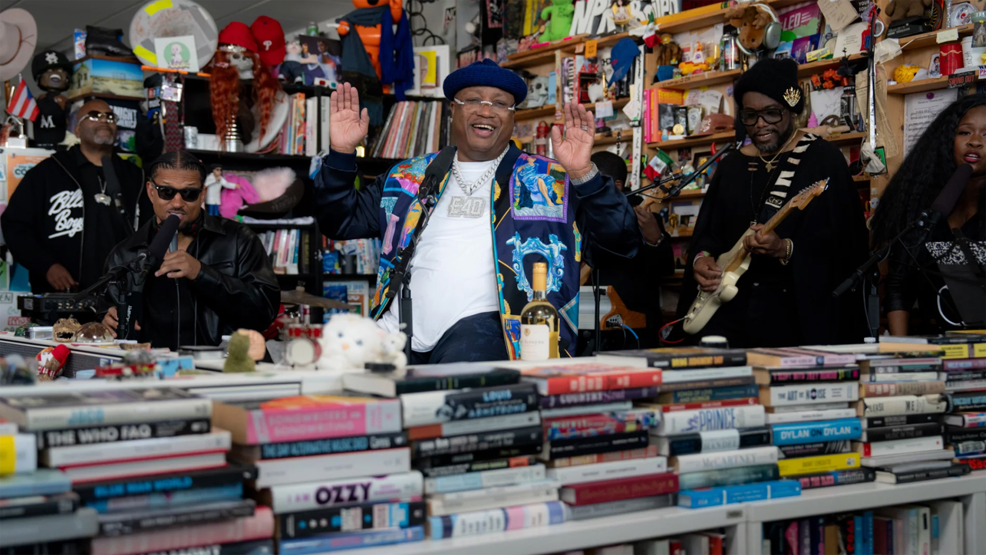 A Black man in glasses and a roll-up beanie smiles and gestures in front of a cluttered bookshelf