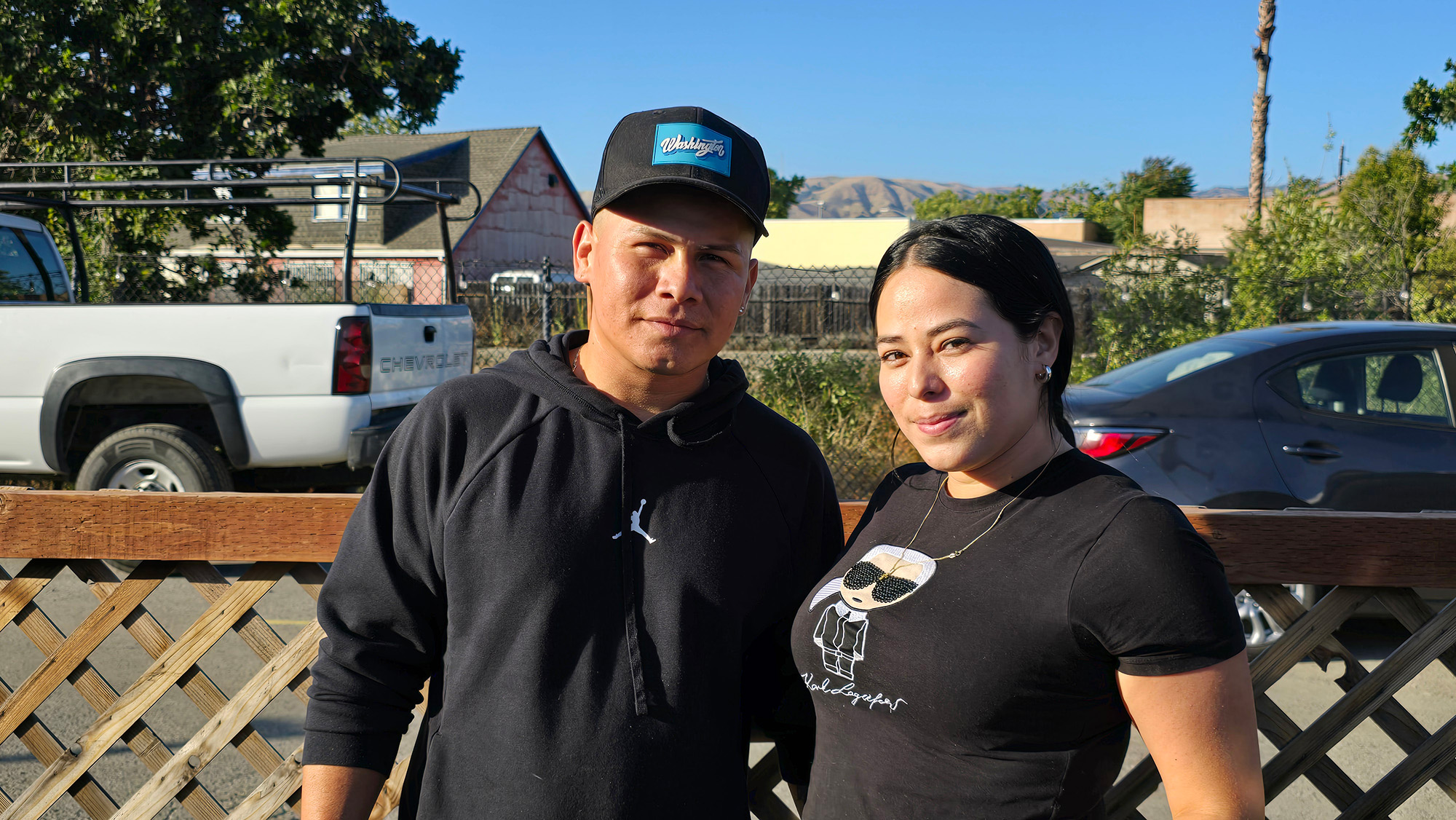 A man and woman, both dressed in black, pose for a portrait next to a fence.