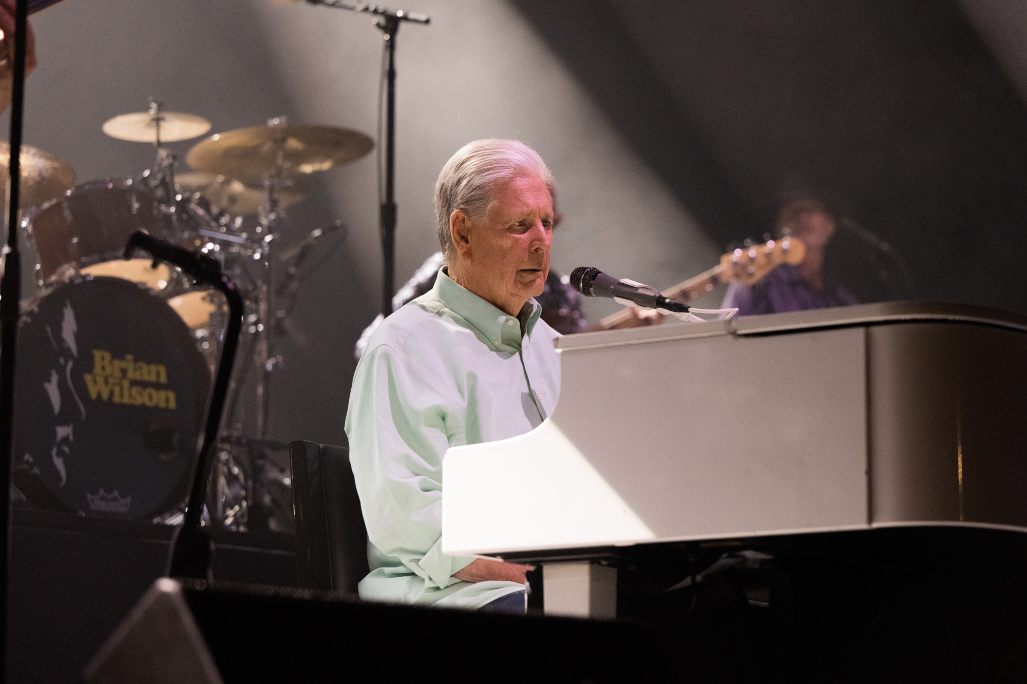 older white man sits at grand piano with mic at face, drum kit in background