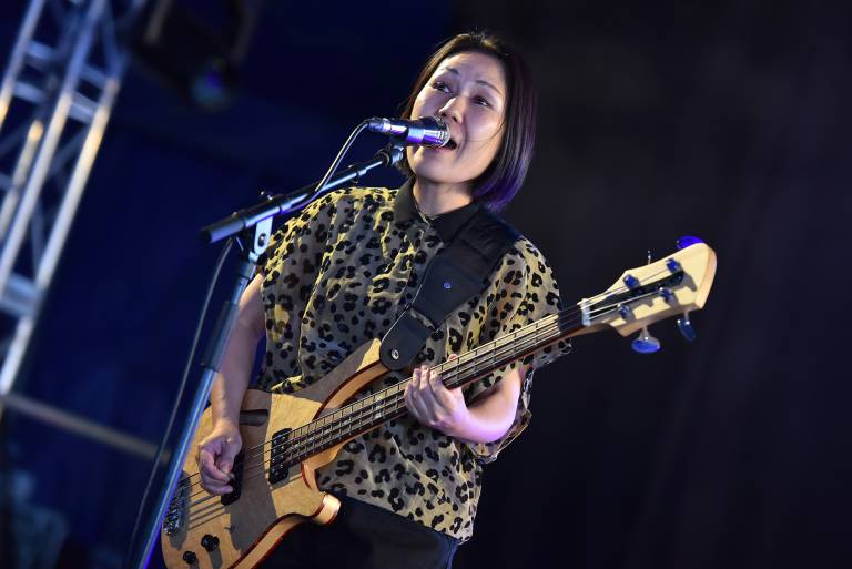 A Japanese American woman plays the bass guitar and sings into a microphone