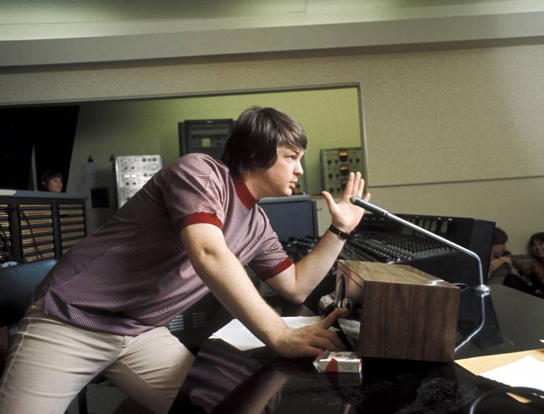 man in T-shirt and white jeans leans over mic in sound booth