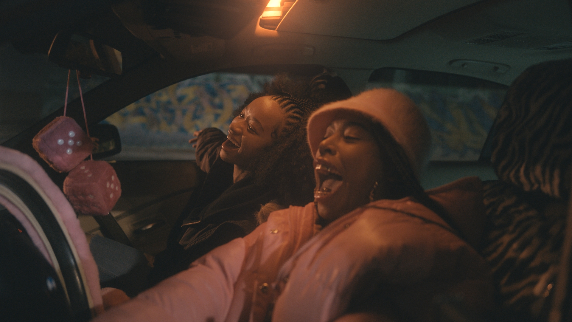 Two people in a car, smiling, on a film set.