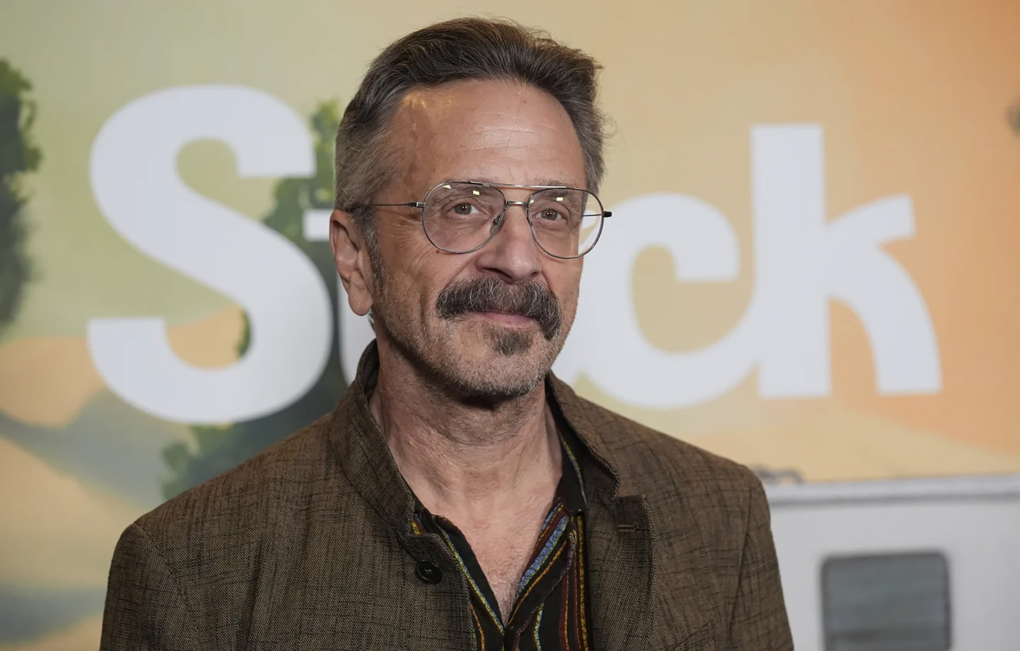 A man in glasses and a mustache poses on a red carpet.