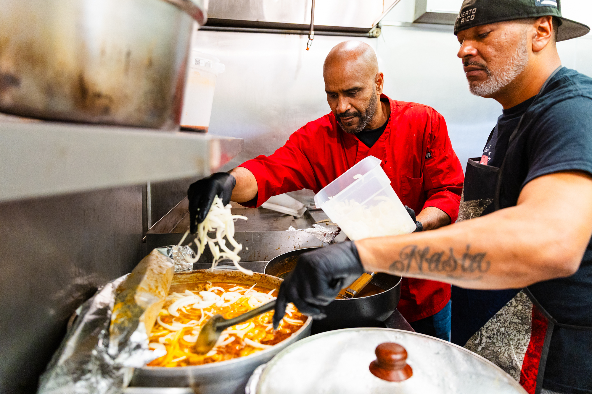 Two chefs add sliced onions to a big pot of stew.