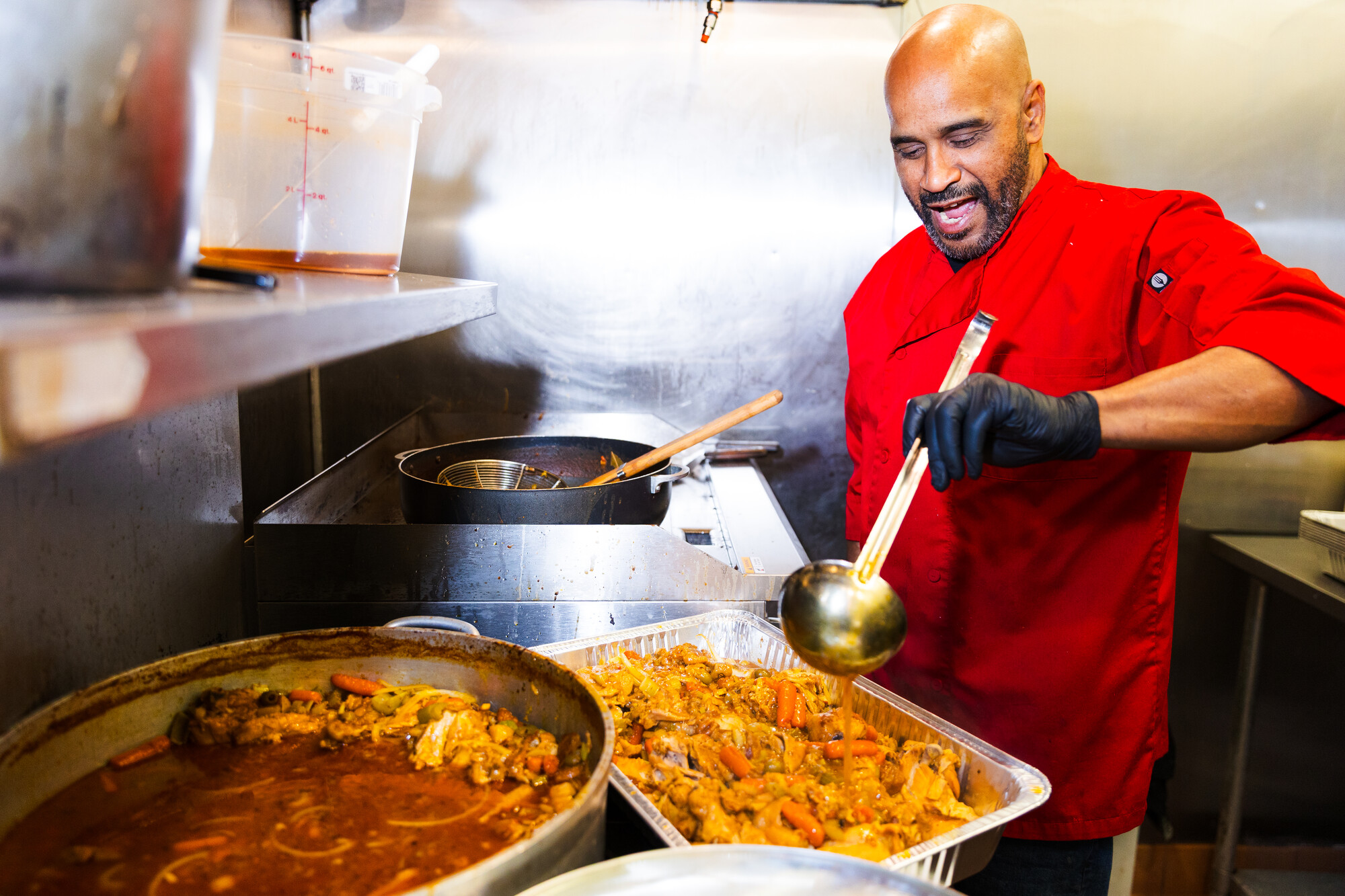 Chef in a red jacket ladles chicken stew into a large aluminum catering tray.
