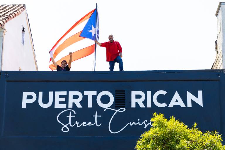 Two men stand on the roof of a restaurant holding a large Puerto Rican flag. The sign on the restaurant reads, "Puerto Rican Street Cuisine."