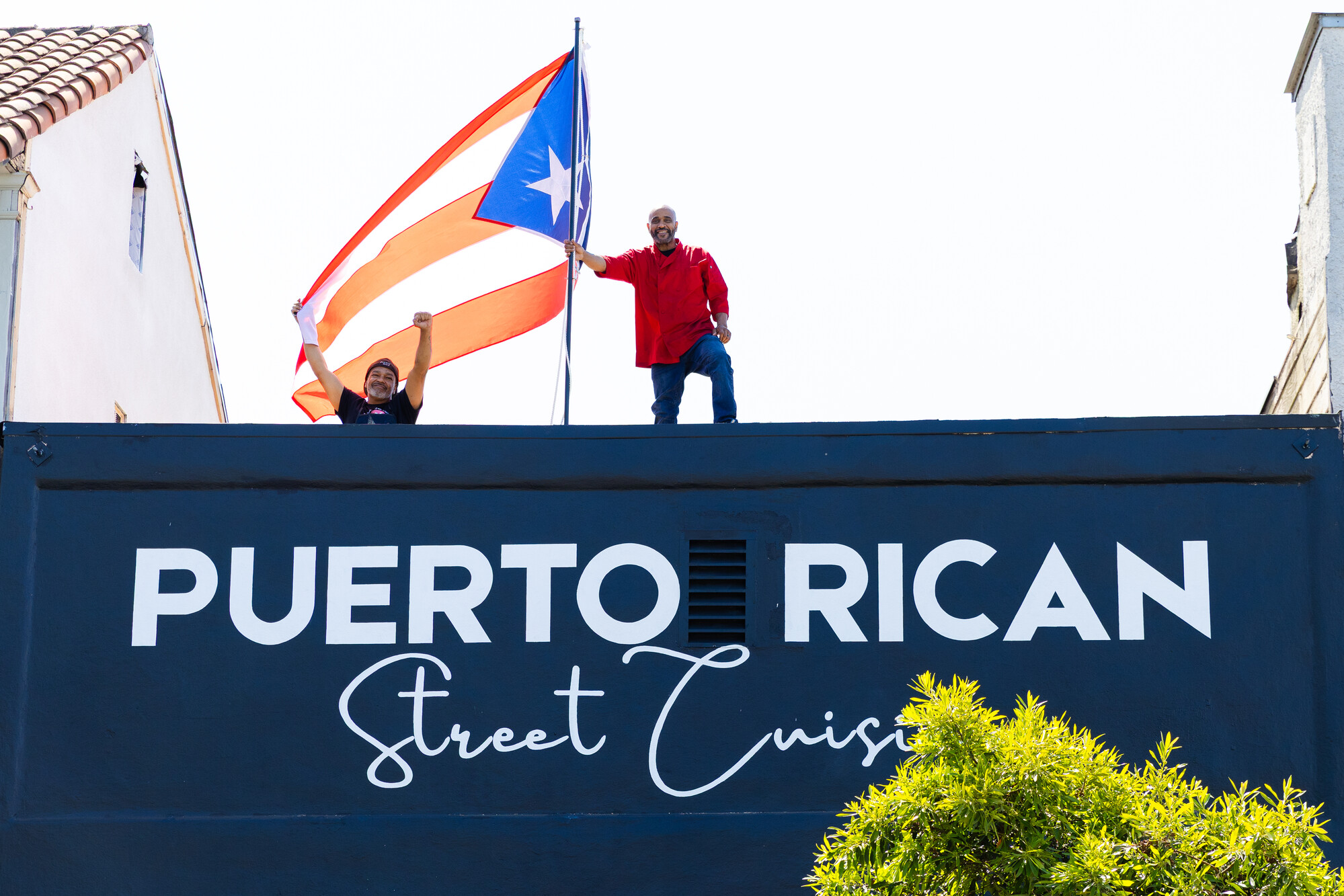 Two men stand on the roof of a restaurant holding a large Puerto Rican flag. The sign on the restaurant reads, "Puerto Rican Street Cuisine."