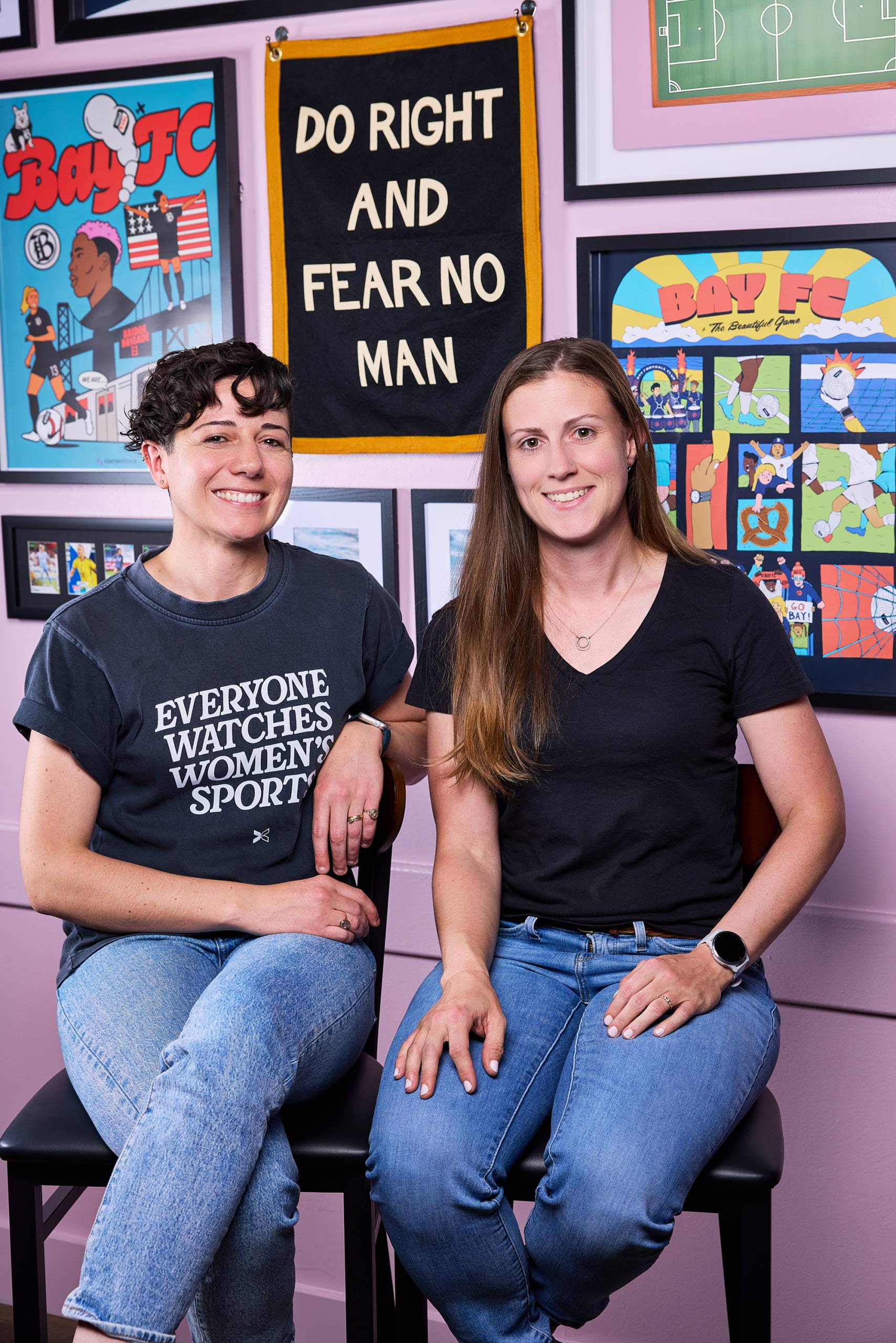 Two women pose for a portrait. The woman on the left wears a black T-shirt that reads, "Everyone watches women's sports."
