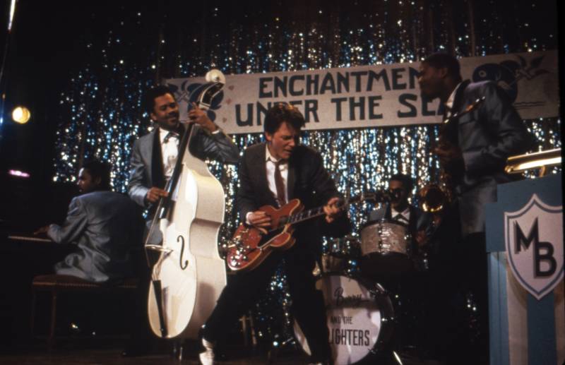 A young white man in a suit and skinny tie plays guitar on a stage decorated with sparkling streamers. Behind him Black men n 1950s style suits play an upright bass, drums, saxophone and piano.