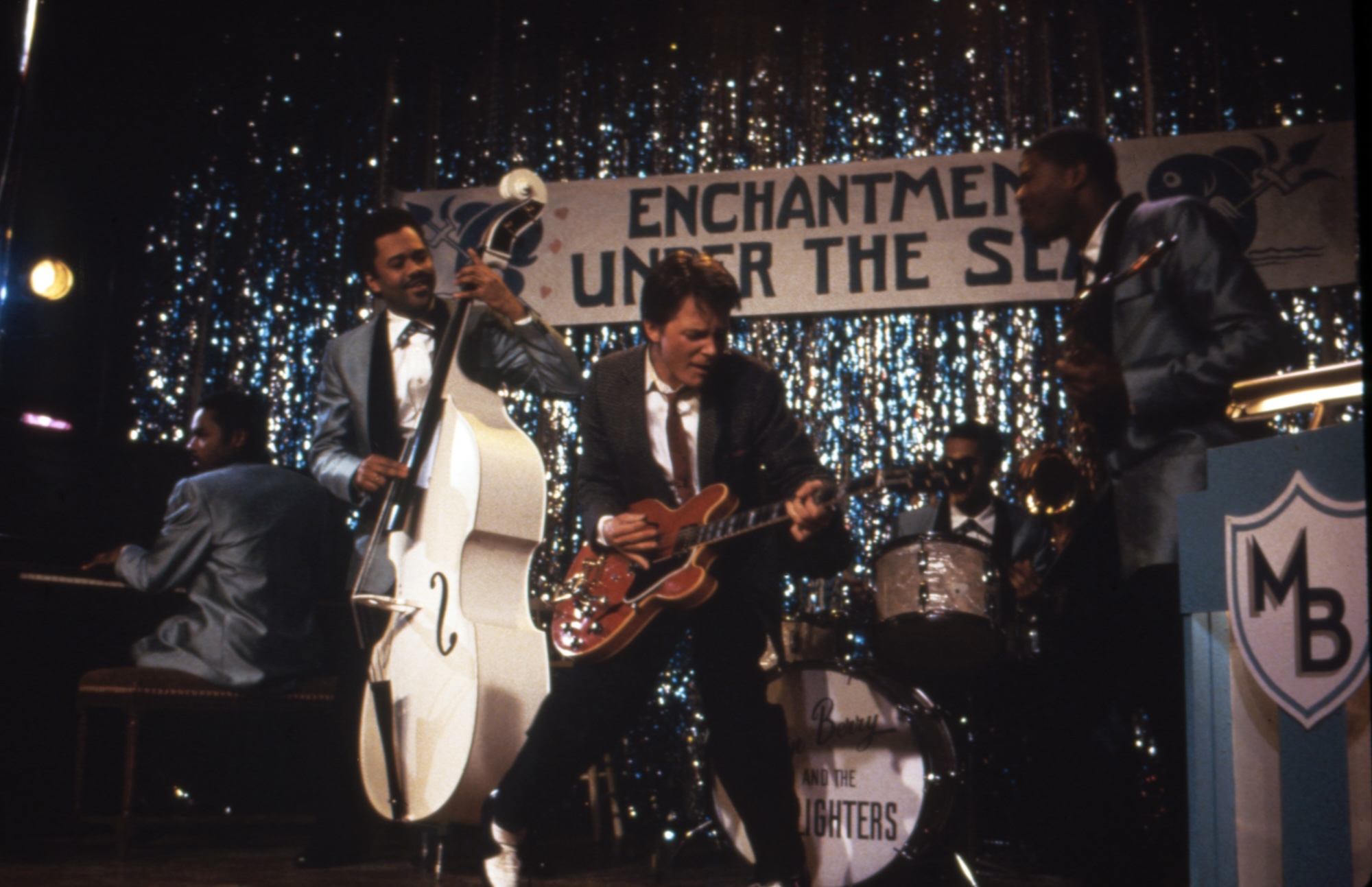 A young white man in a suit and skinny tie plays guitar on a stage decorated with sparkling streamers. Behind him Black men n 1950s style suits play an upright bass, drums, saxophone and piano.